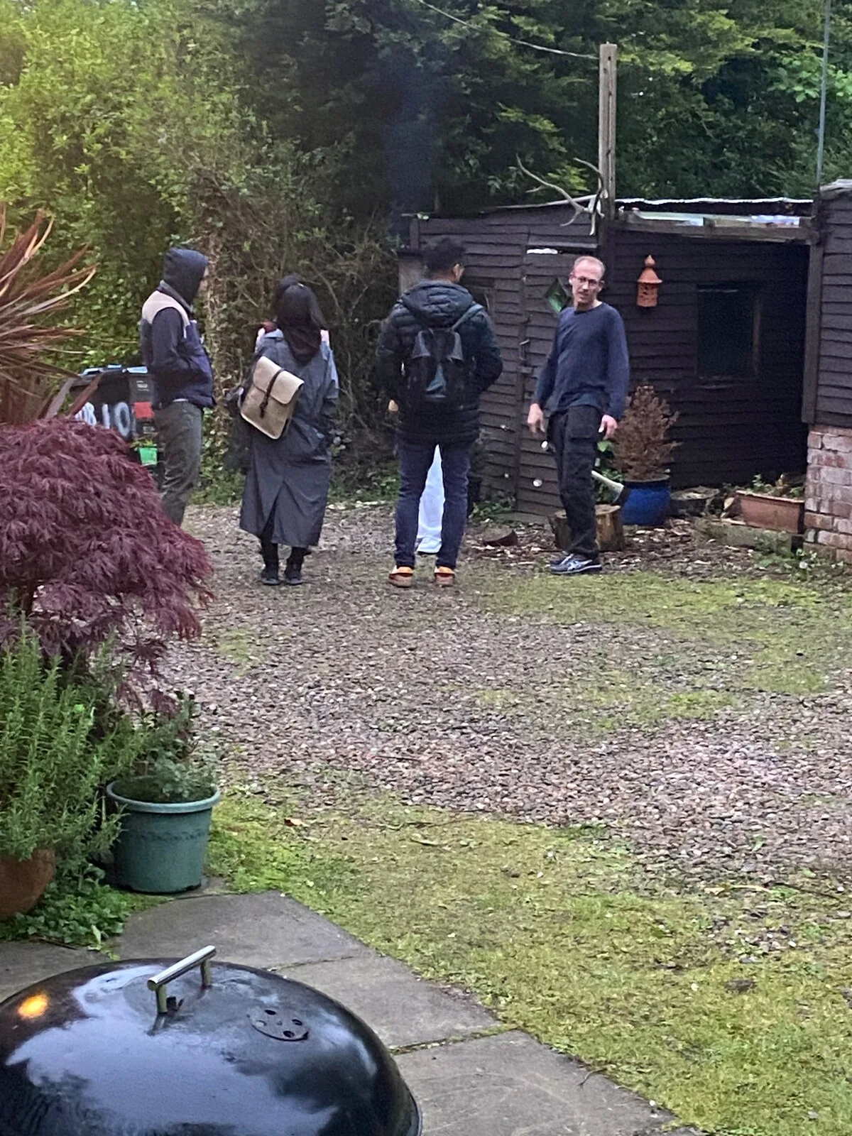 People stand in a gravel garden beside a dark wooden shed.