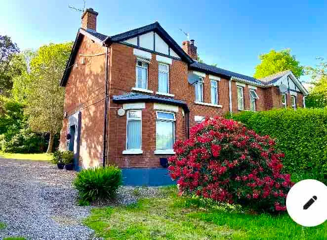 A red-brick house stands beside a gravel driveway and flowering shrubs.