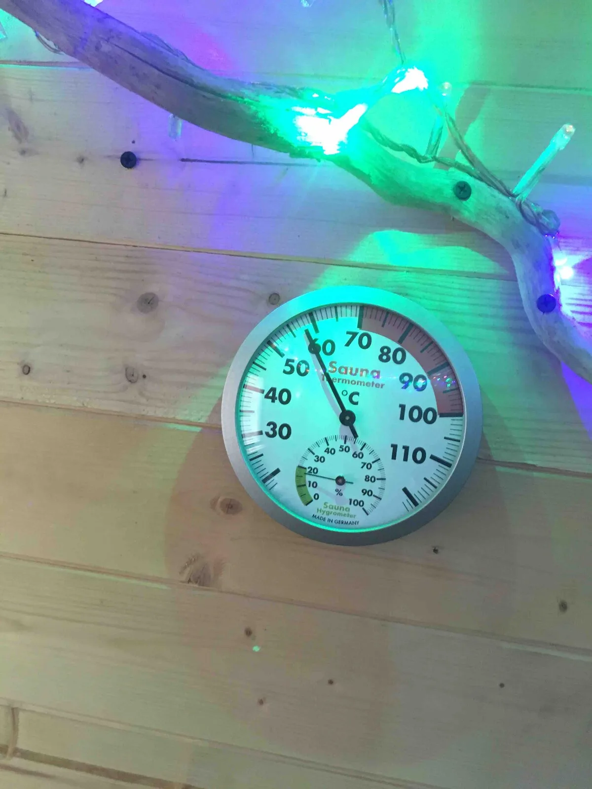 A round thermometer is mounted on a wooden wall beneath colorful lights.