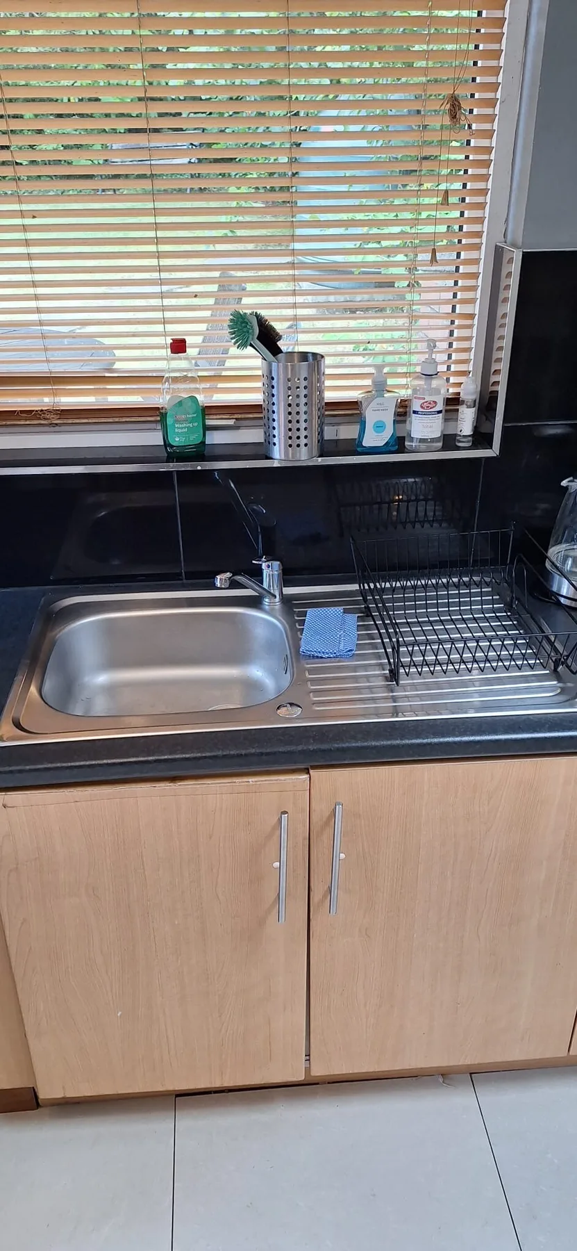 A kitchen sink and dish rack sit beneath a window with blinds.