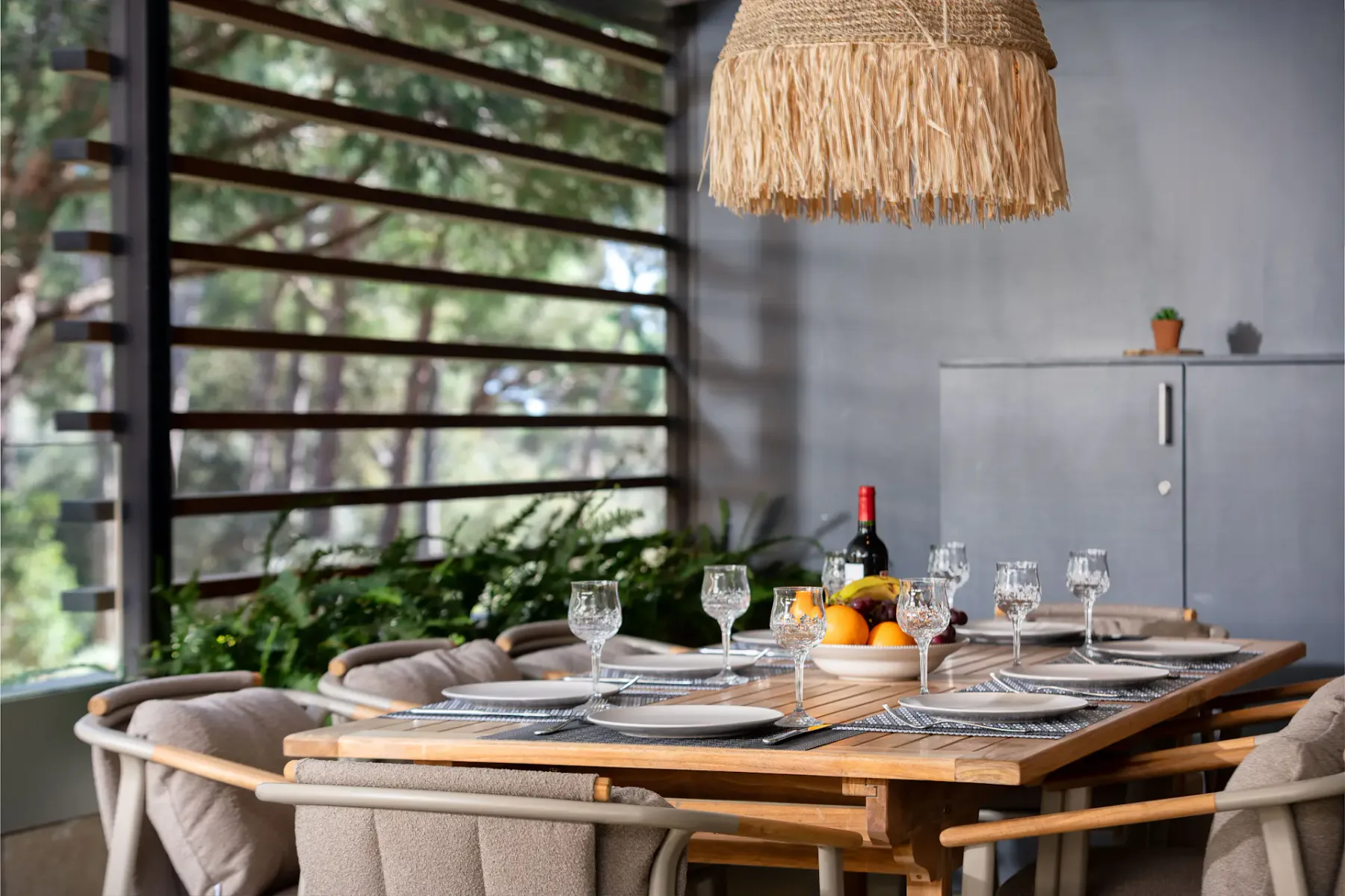 A dining area with a set table beside large windows and leafy plants.