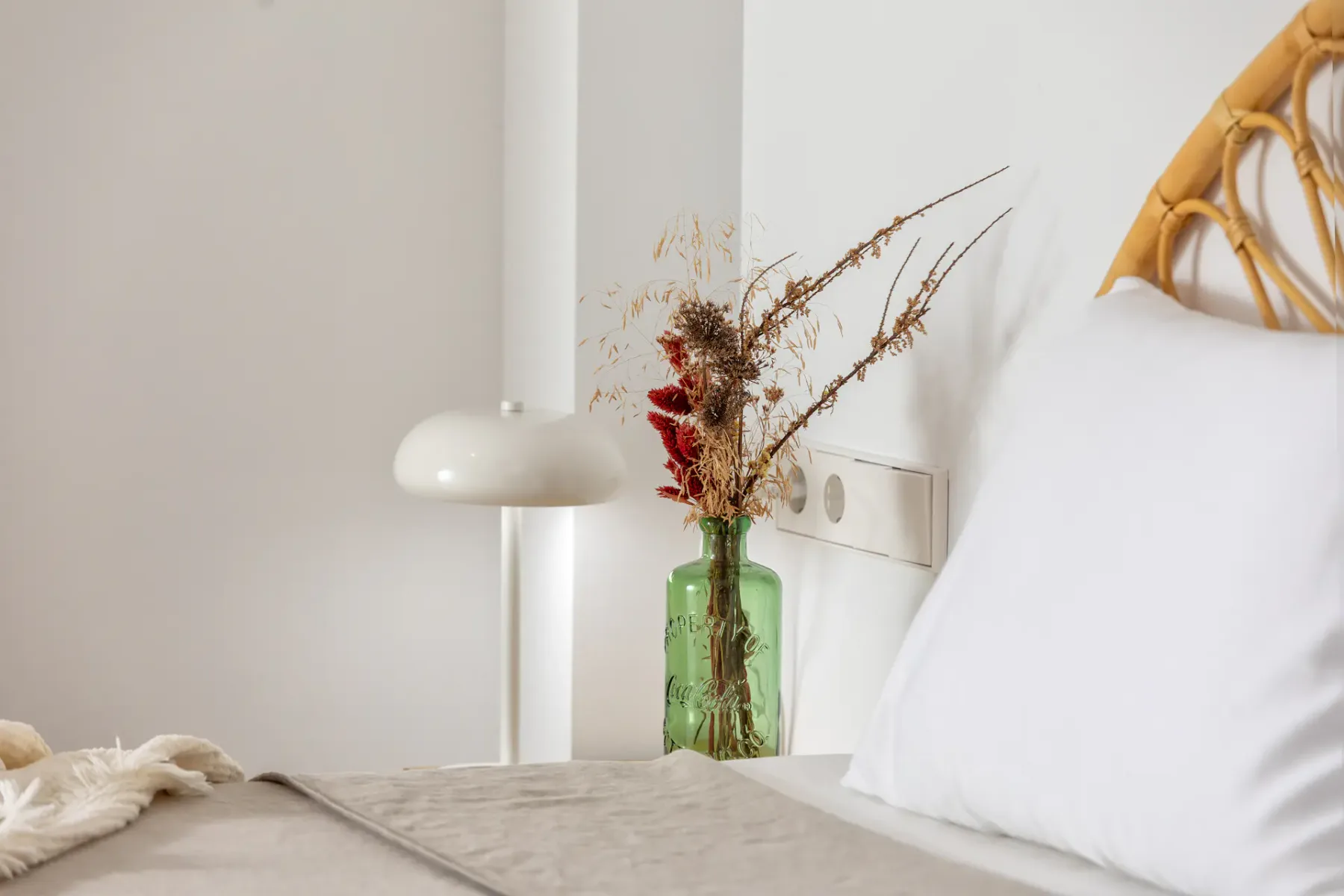 A bedroom with a bed, white lamp, and vase of dried flowers.