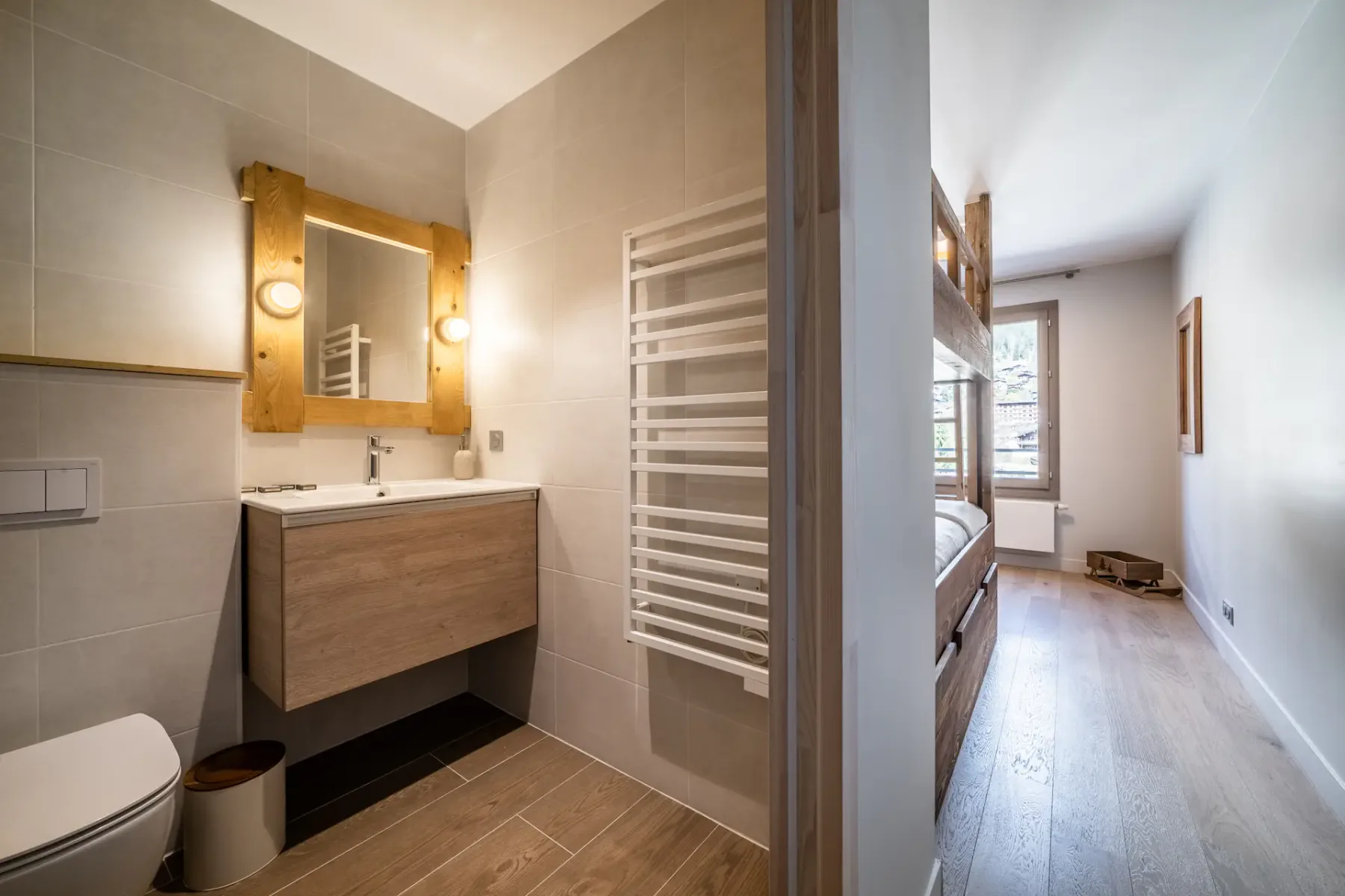 Un baño con mueble de lavabo de frente de madera, espejo de pared y radiador toallero junto a un pasillo.