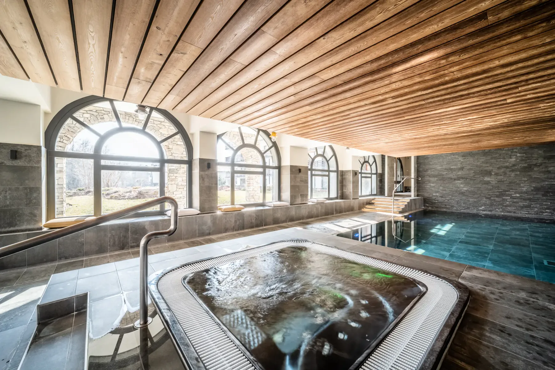 Una piscina interior y un spa con burbujas están bajo un techo revestido de madera.