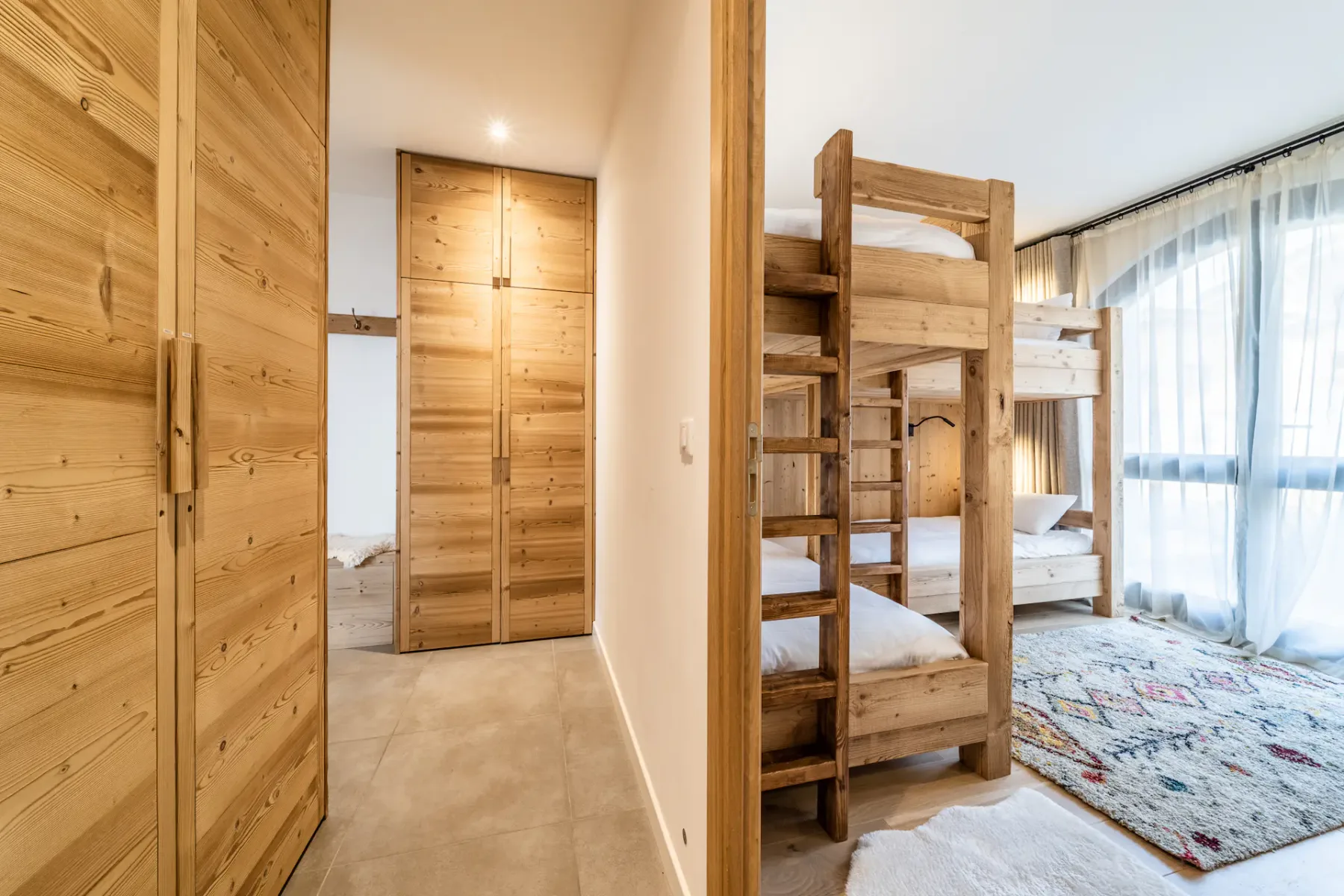 Un dormitorio con literas de madera junto a una ventana luminosa y un pasillo con armarios de madera.