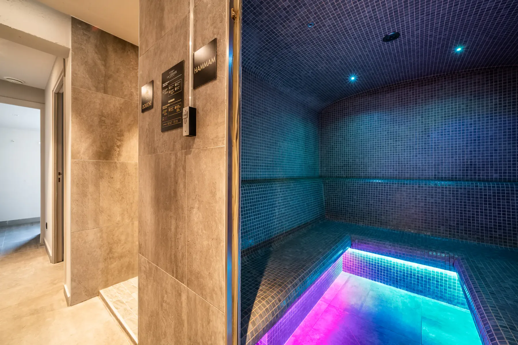 Una piscina interior revestida de azulejos brilla con luces azules y rosas junto a una entrada de piedra.