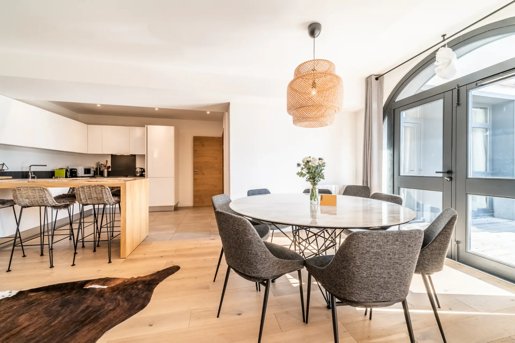 Una luminosa zona de comedor y cocina con una mesa redonda junto a grandes ventanas.