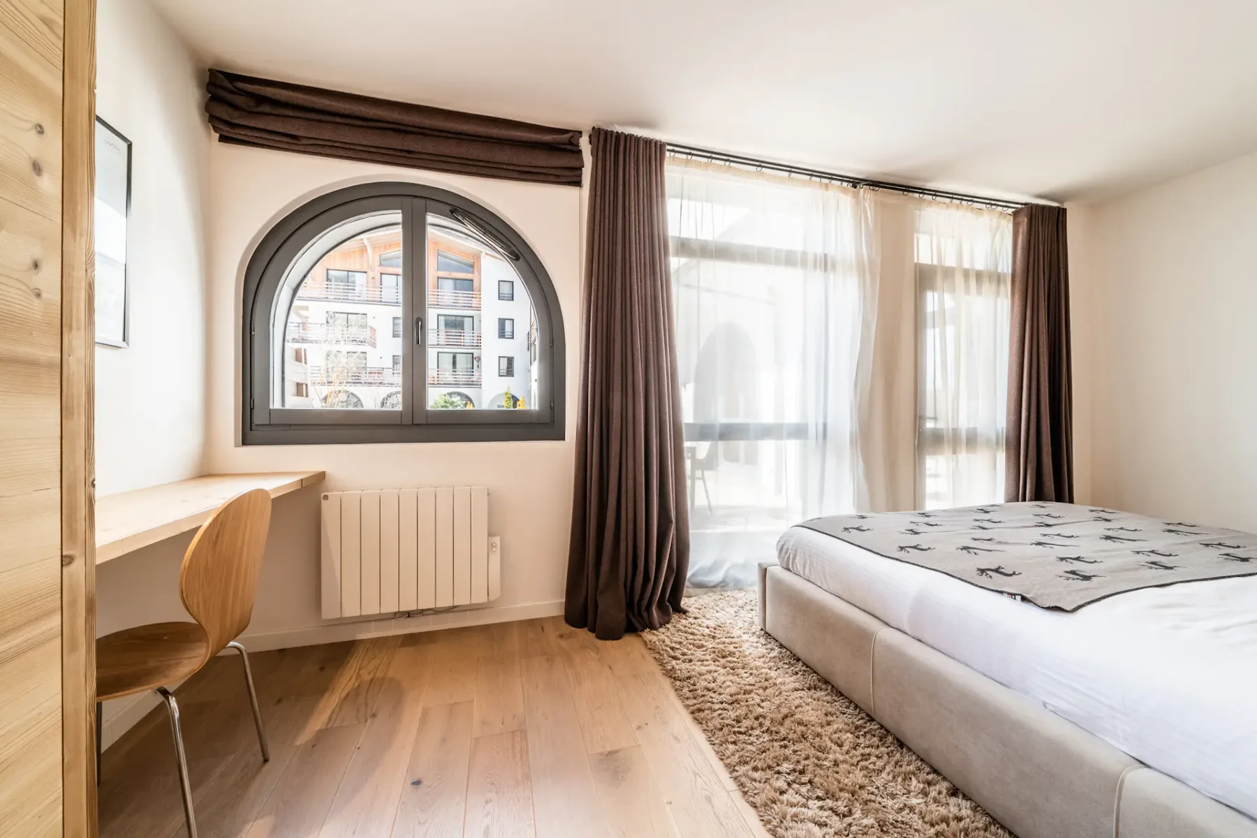 Un dormitorio con cama, escritorio y ventana arqueada junto a grandes ventanales con cortinas.