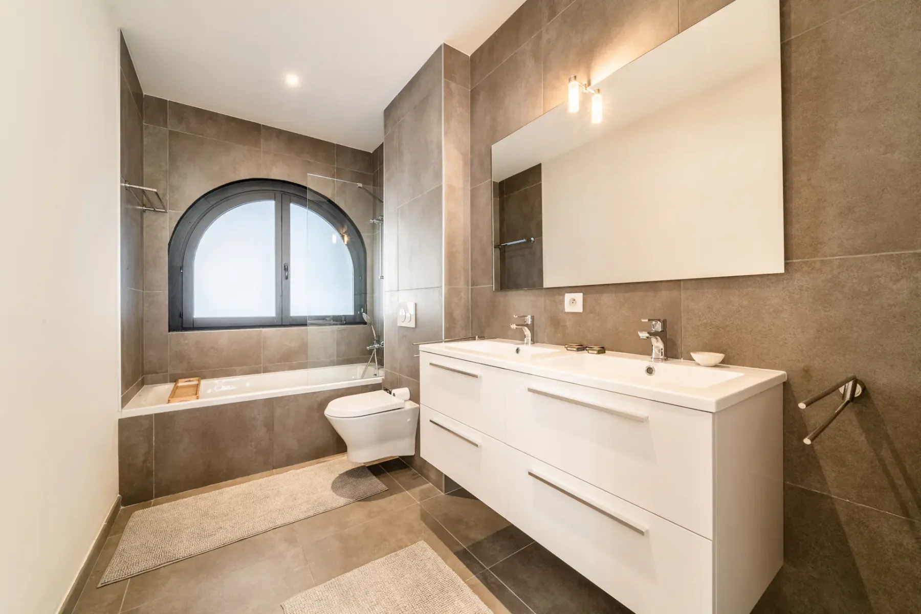 Un baño moderno con bañera bajo una ventana arqueada y un tocador blanco.