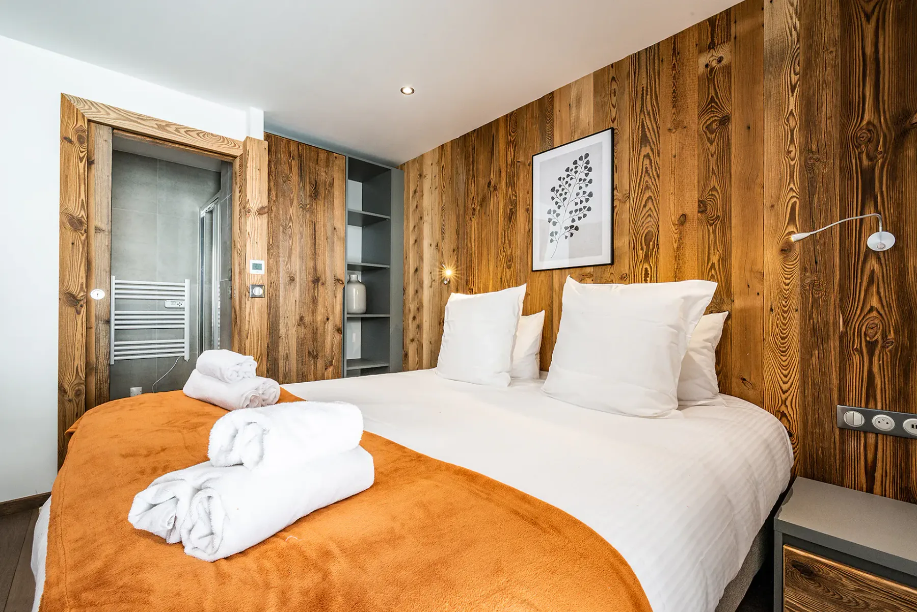 Un dormitorio con una cama blanca, una manta naranja y paredes revestidas de madera.
