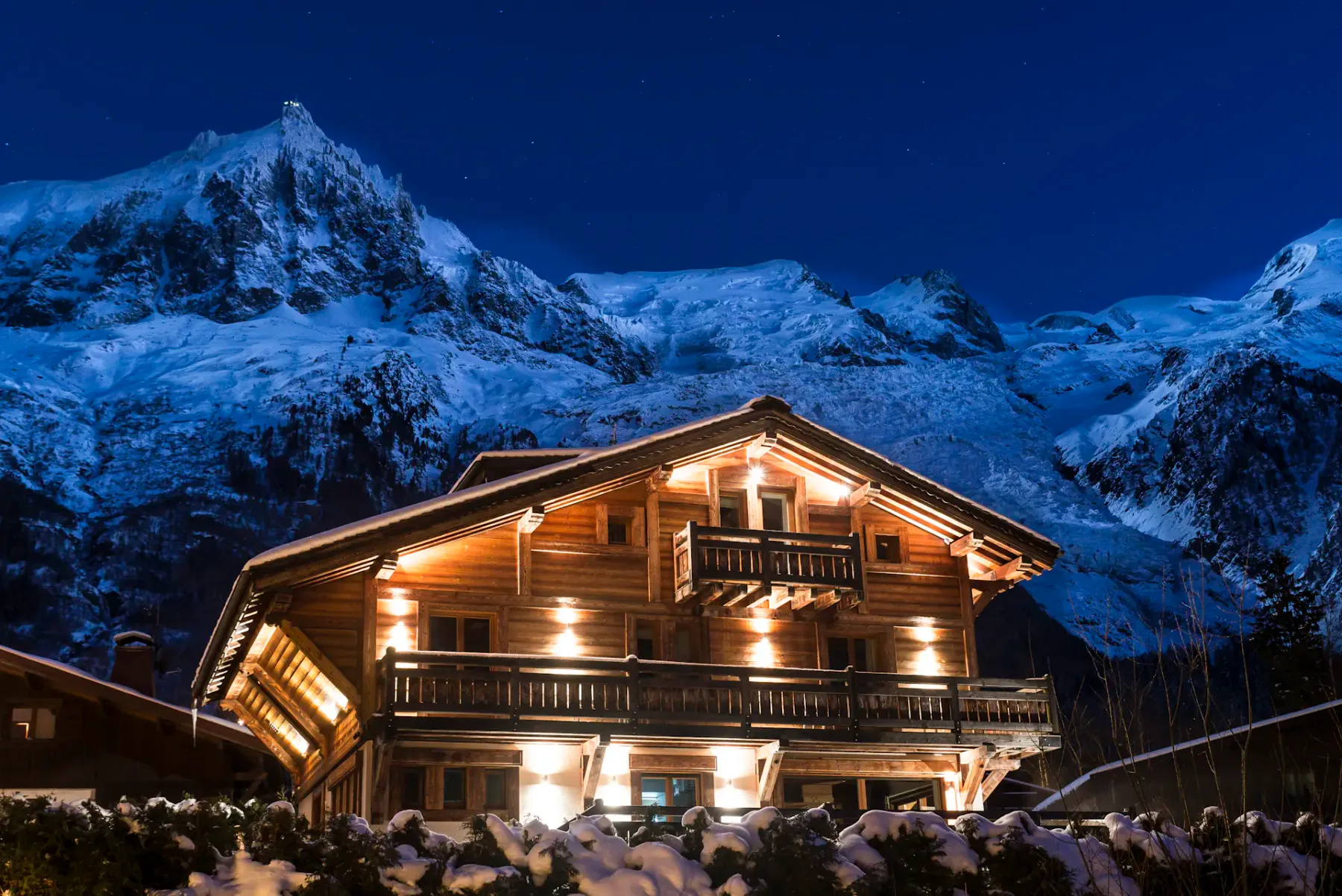 Un chalet de montaña de madera iluminado se alza bajo picos nevados por la noche.