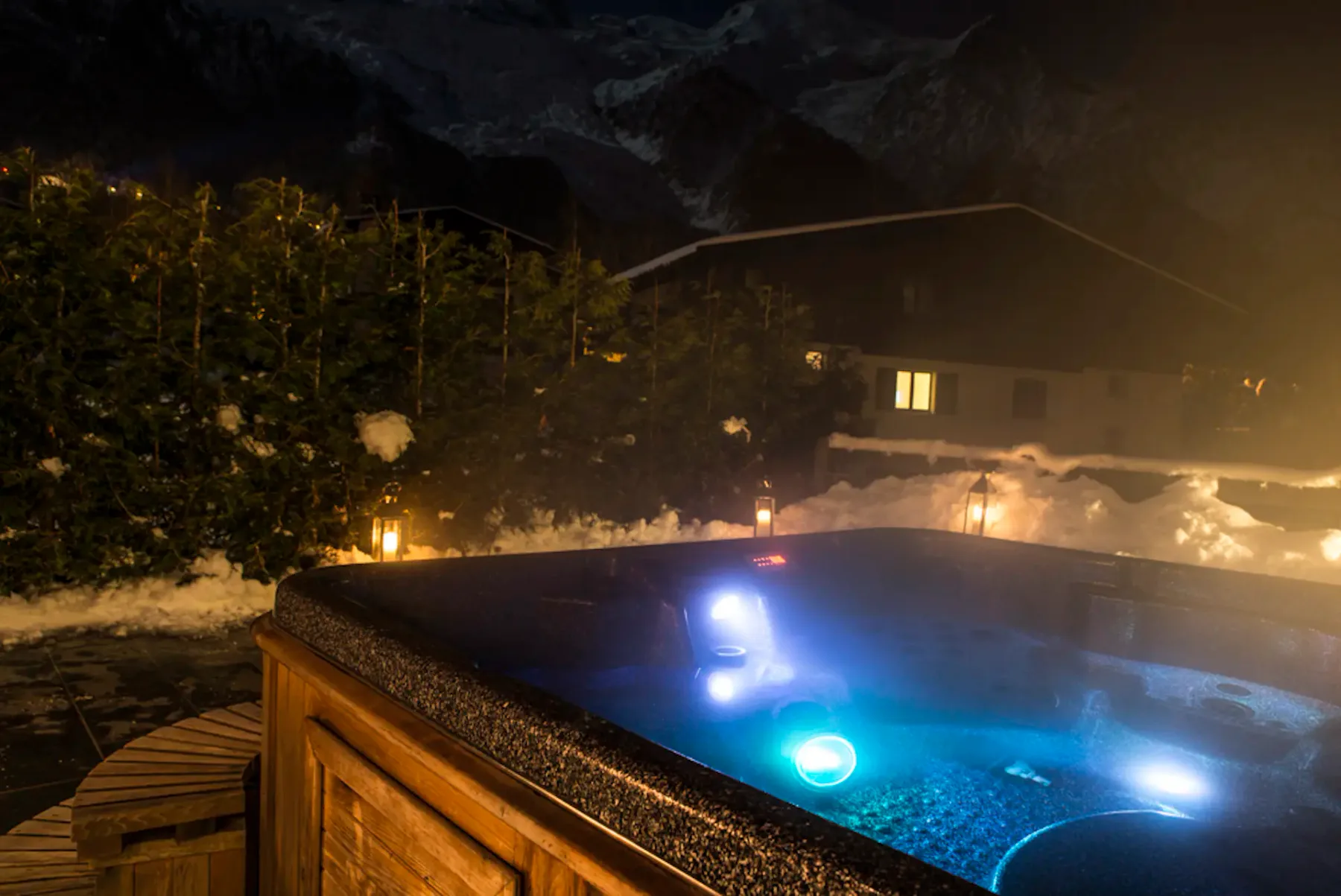Una bañera de hidromasaje exterior brilla junto a un alojamiento de montaña cubierto de nieve por la noche.