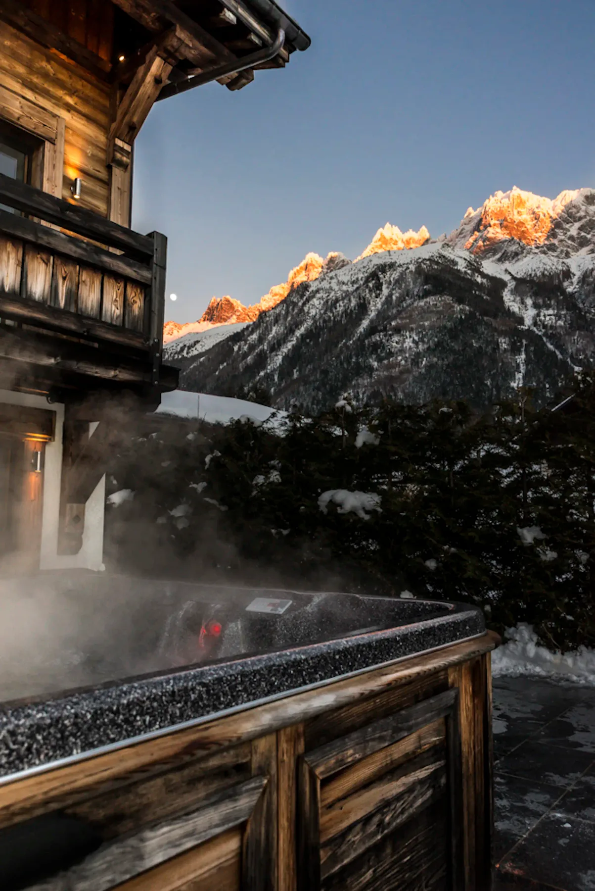 Una bañera de hidromasaje exterior humeante está junto a un edificio de madera, con montañas nevadas al fondo.