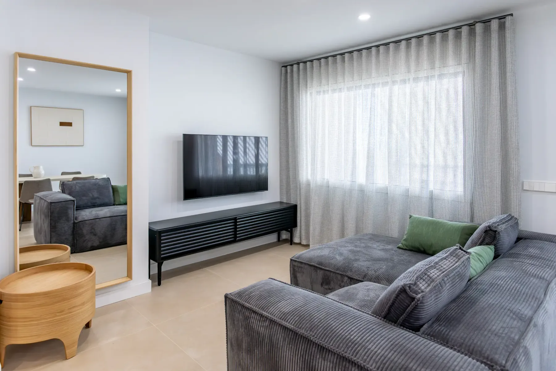 A living room with gray seating, a wall-mounted television, and a large curtained window.