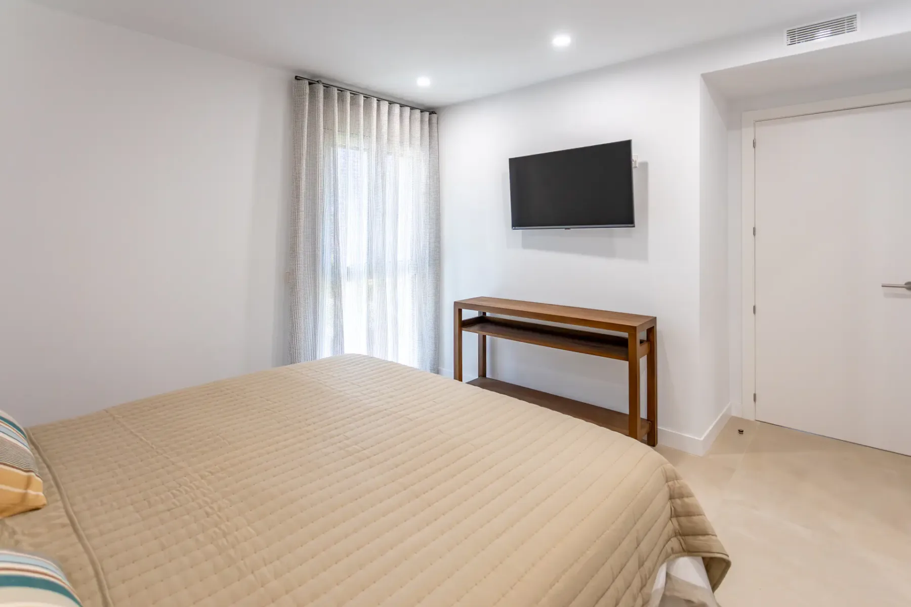 A bright bedroom with a bed, a wall-mounted television, and a wooden table.