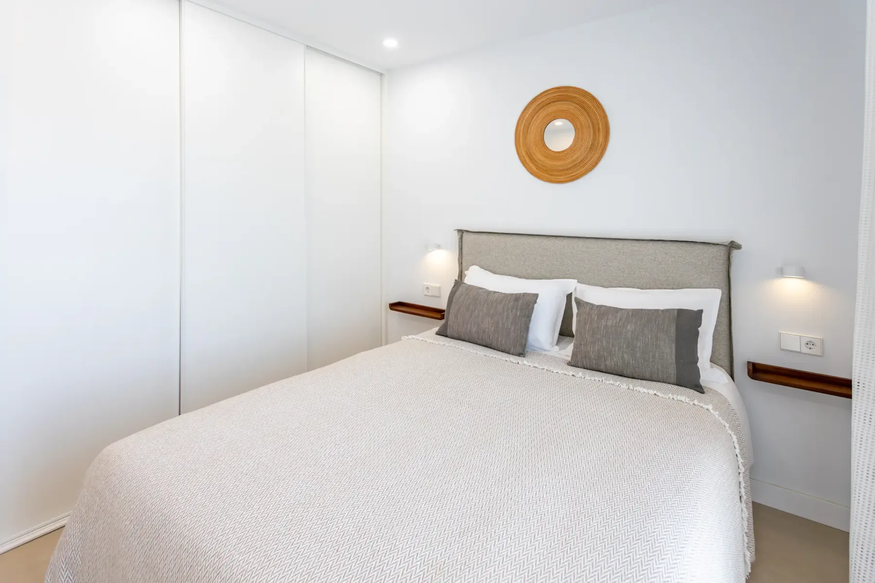 A bright bedroom with a neatly made bed and round wall mirror.