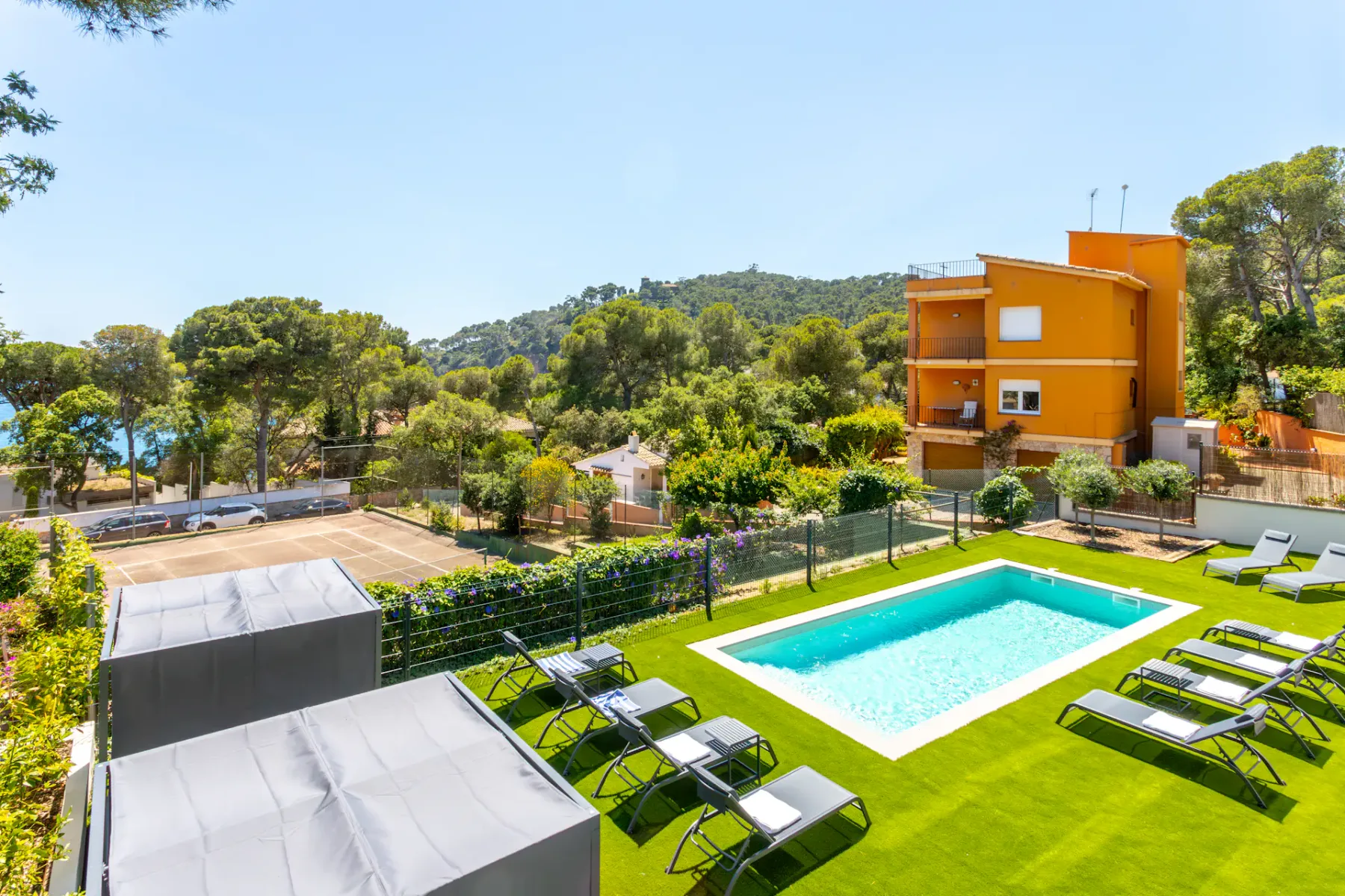An outdoor swimming pool and sun loungers sit beside an orange building among trees.