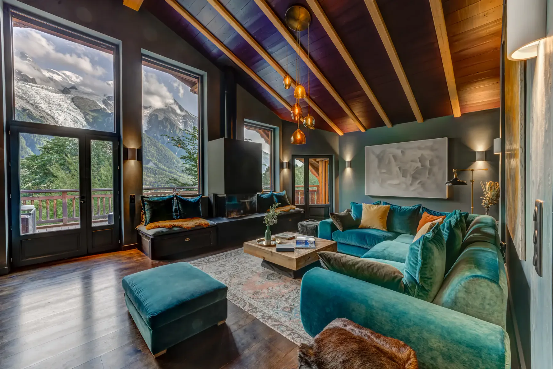 A living room has a teal sectional, a low coffee table, and tall windows overlooking a mountain landscape.