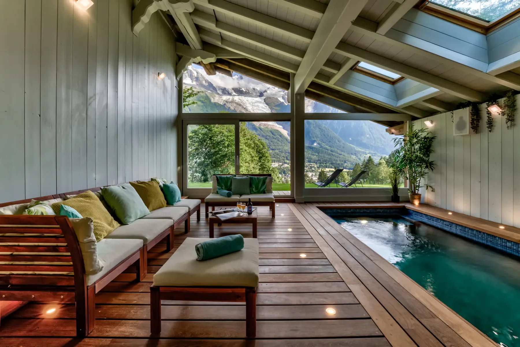 Indoor pool lounge with cushioned seating, a wooden deck, and large windows overlooking mountain scenery.