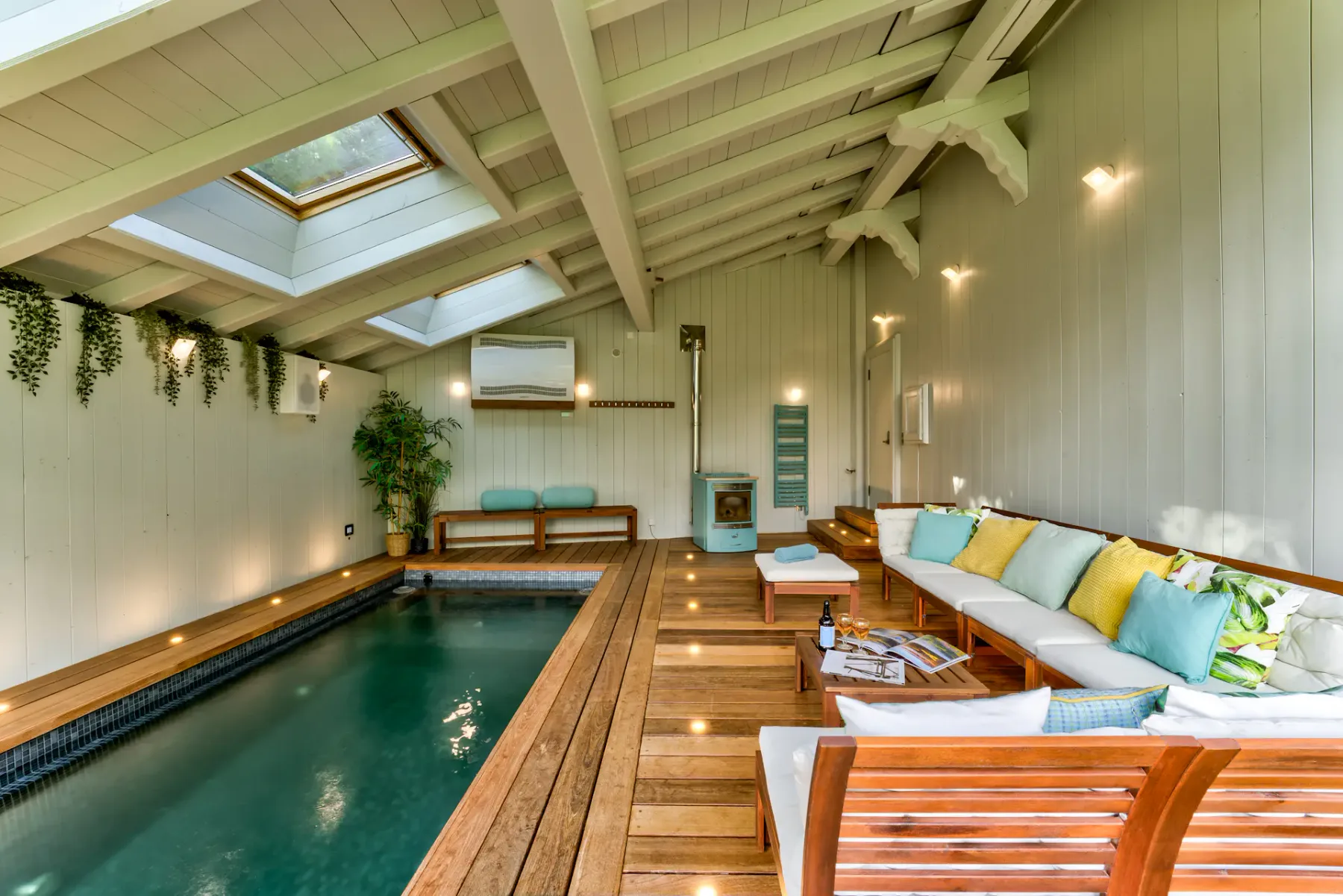 A compact indoor pool sits beside a wooden deck with lounge seating beneath a bright, beamed ceiling.