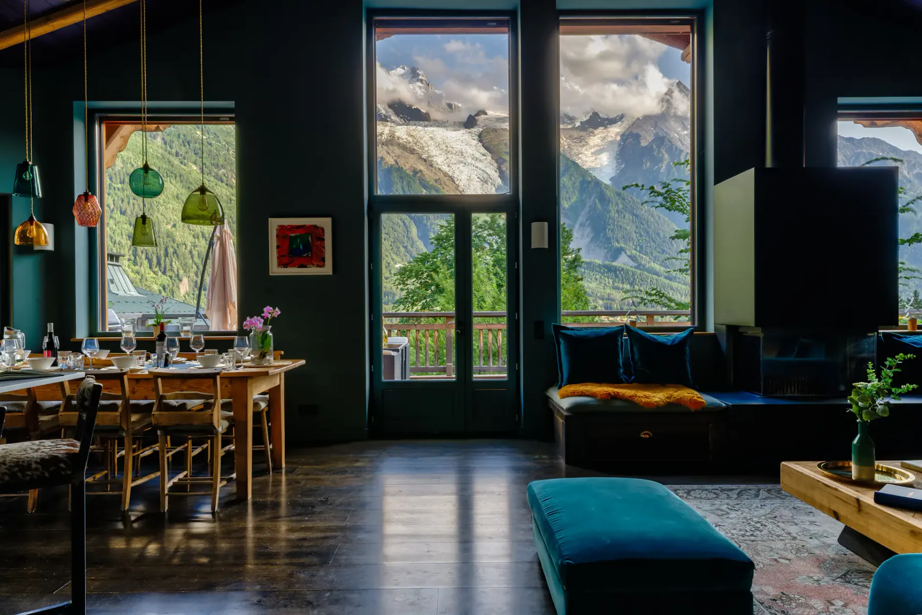 A dining table and lounge area sit beside tall windows overlooking a mountain landscape.
