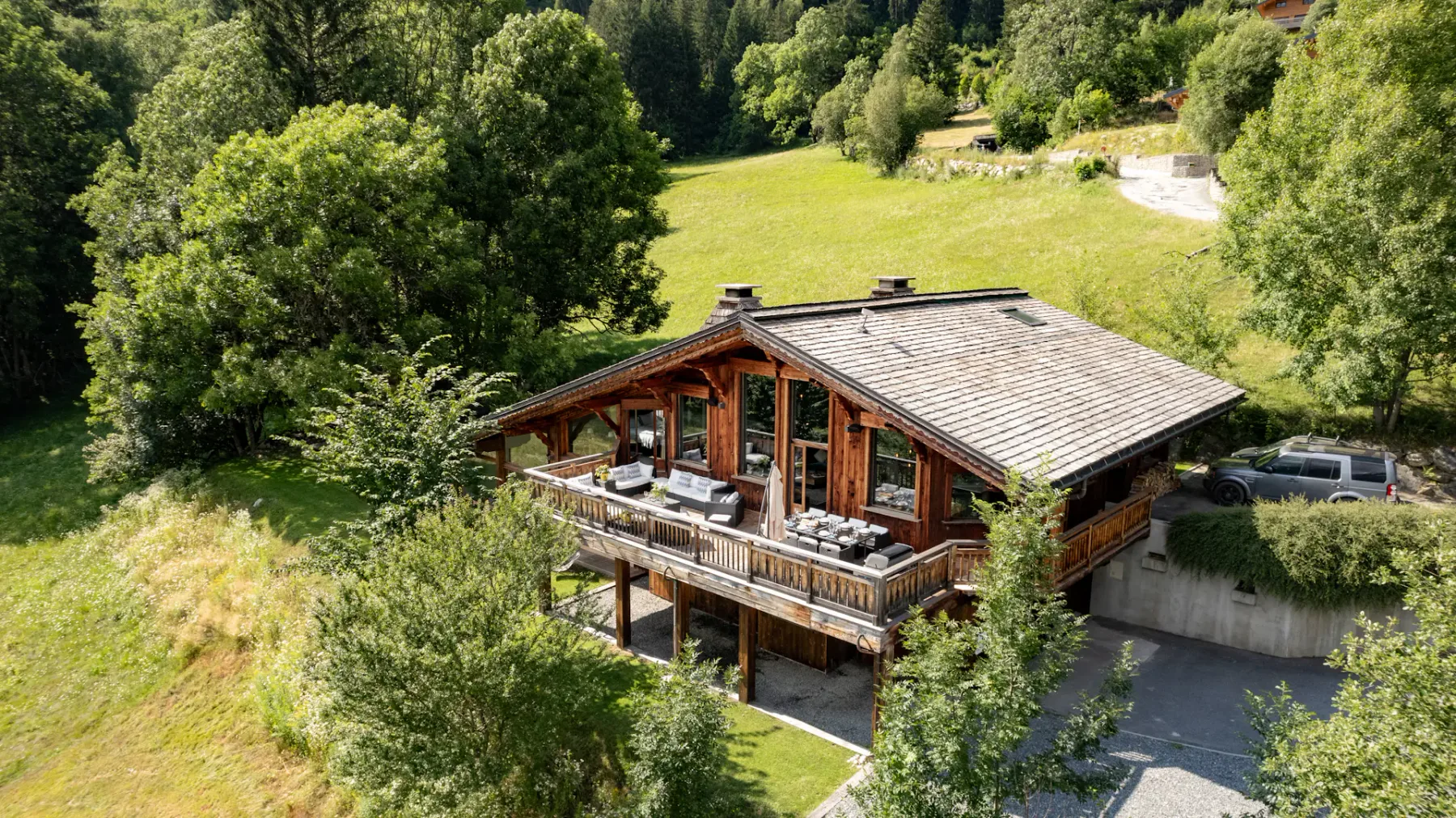 A wooden chalet with a broad roof and furnished balcony sits beside a driveway in a green, wooded landscape.