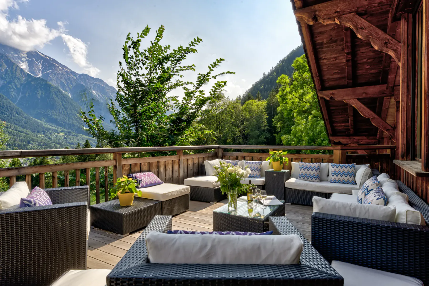A wooden mountain-view terrace features wicker seating, patterned cushions, potted plants, and a glass coffee table.