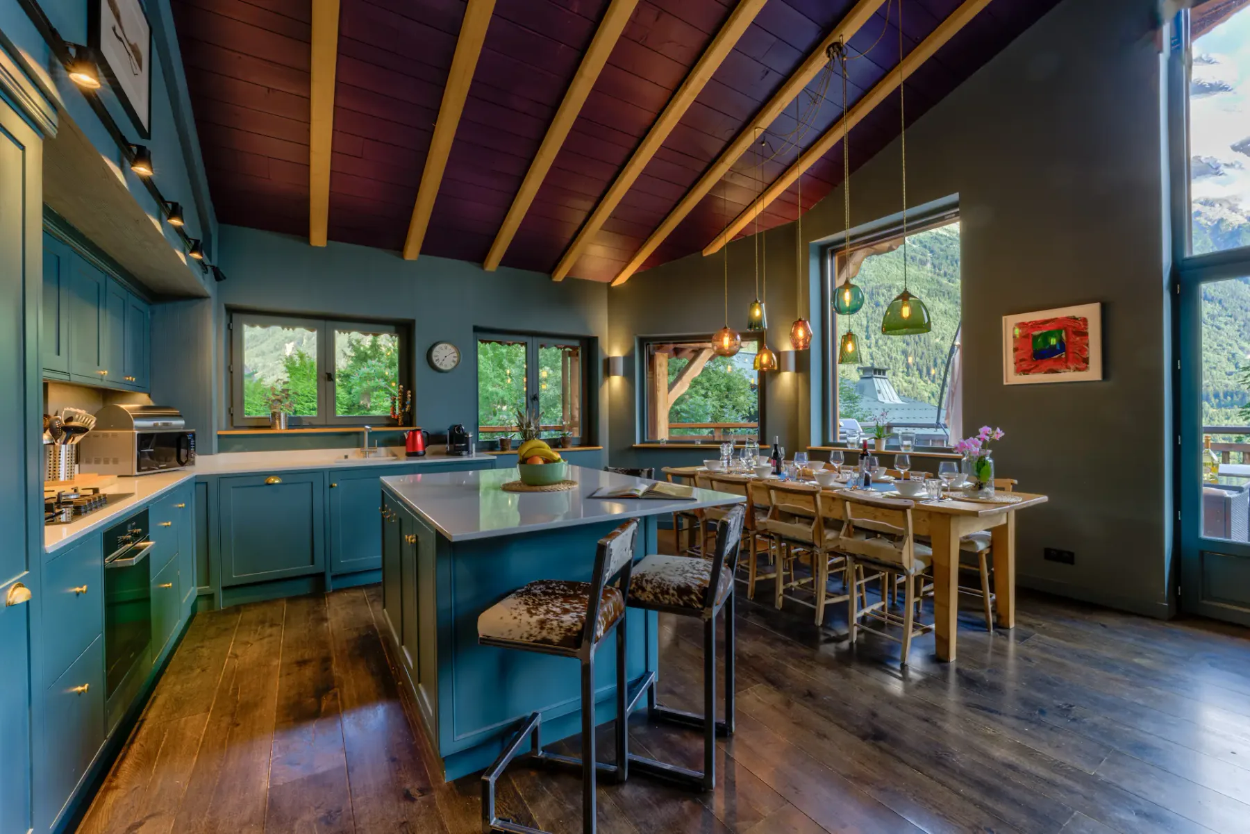 A spacious kitchen with teal cabinets, a central island, and a long dining table beneath exposed wooden beams.