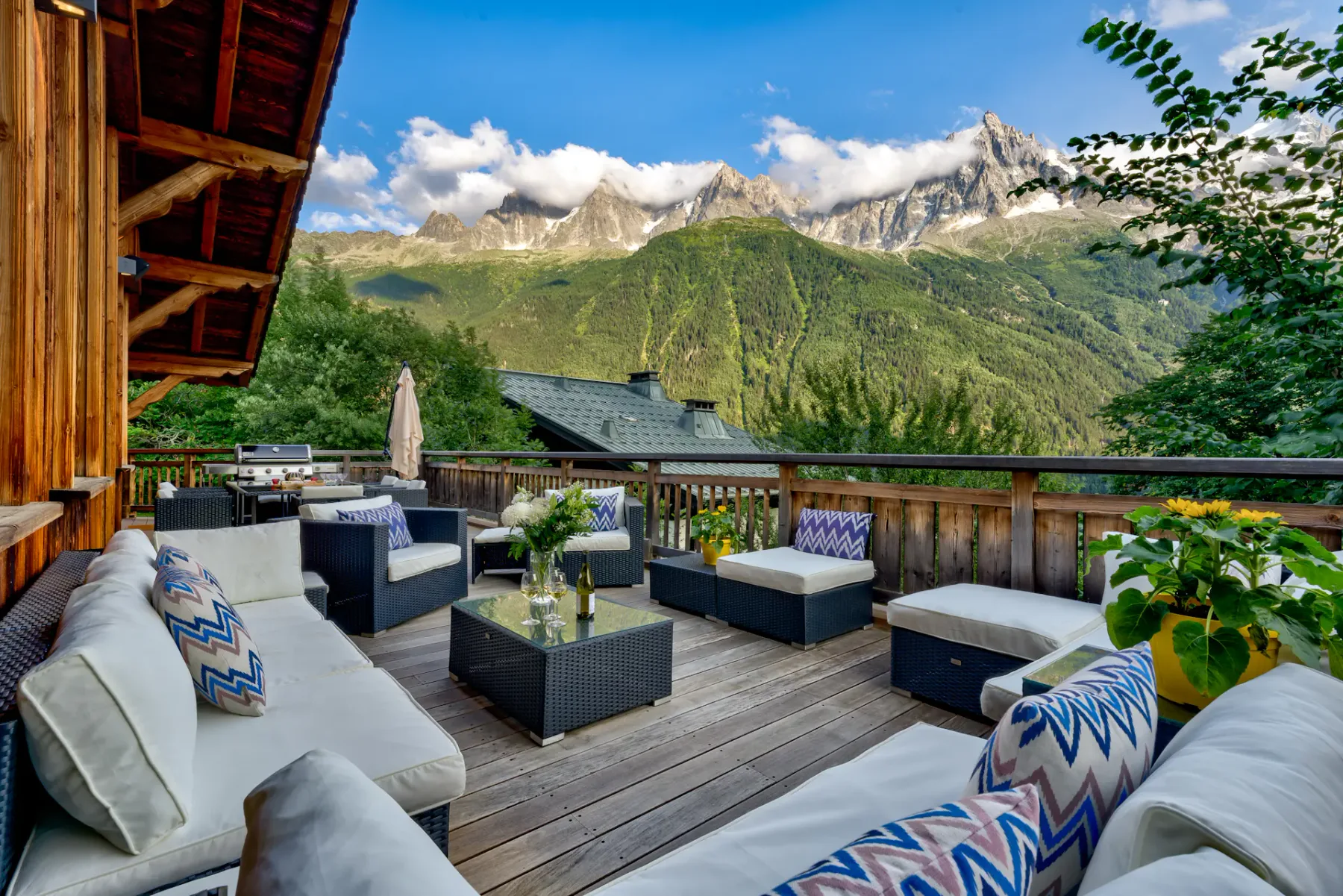 A wooden deck has cushioned outdoor seating and a table overlooking green mountains and rocky peaks.