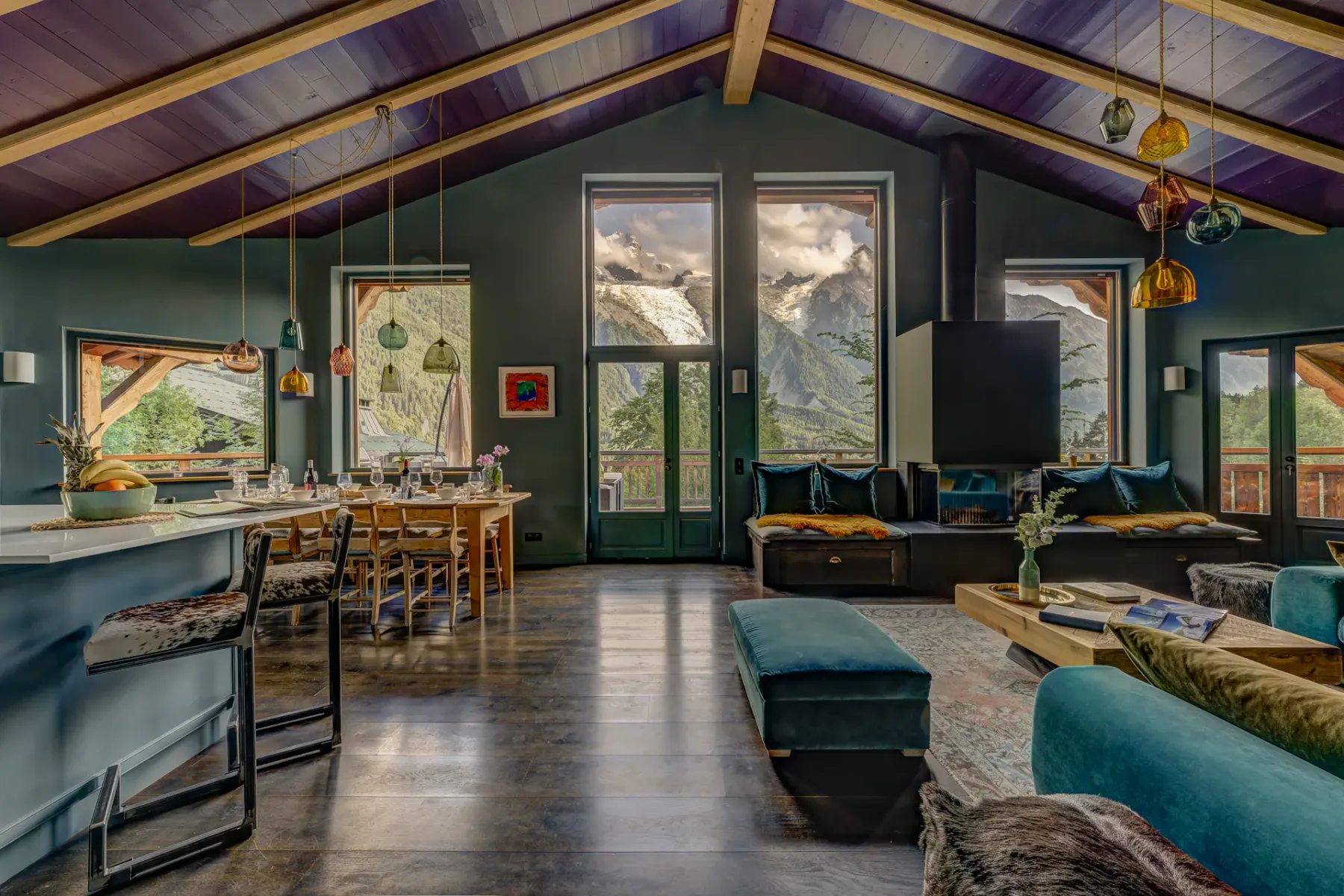 A vaulted living room has exposed timber beams, large mountain-view windows, and teal seating around a dining table.