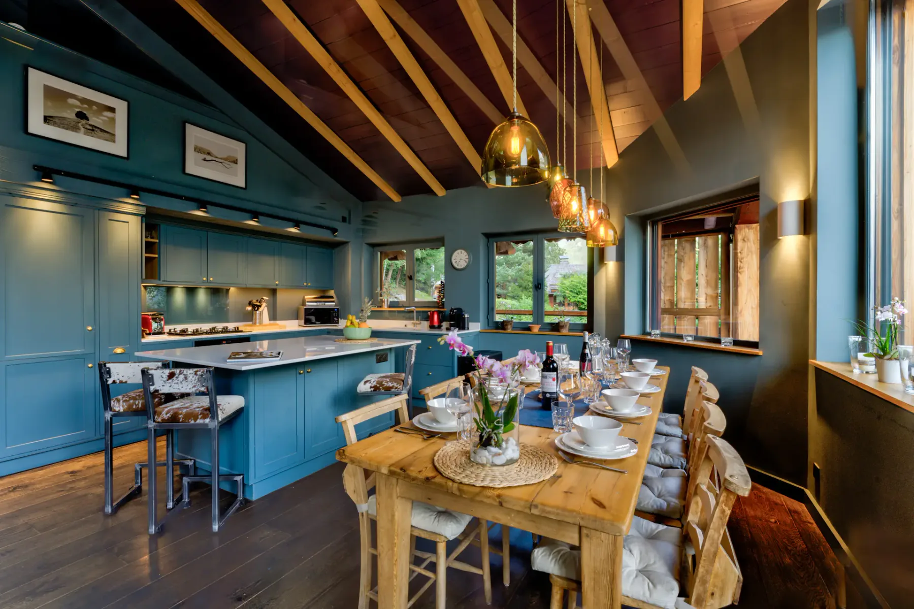 A dining table is set with dishes beneath pendant lights in a blue kitchen with large windows.