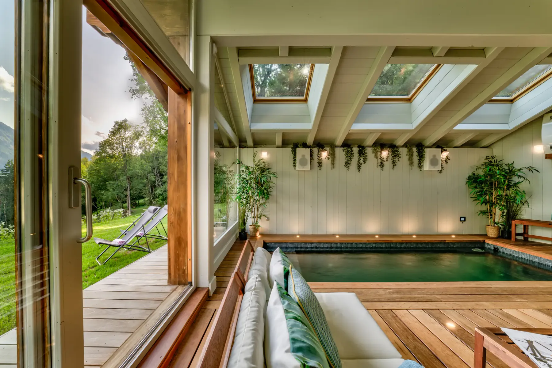 An indoor pool sits beneath skylights, with lounge seating, potted plants, and a glass door opening onto a deck.