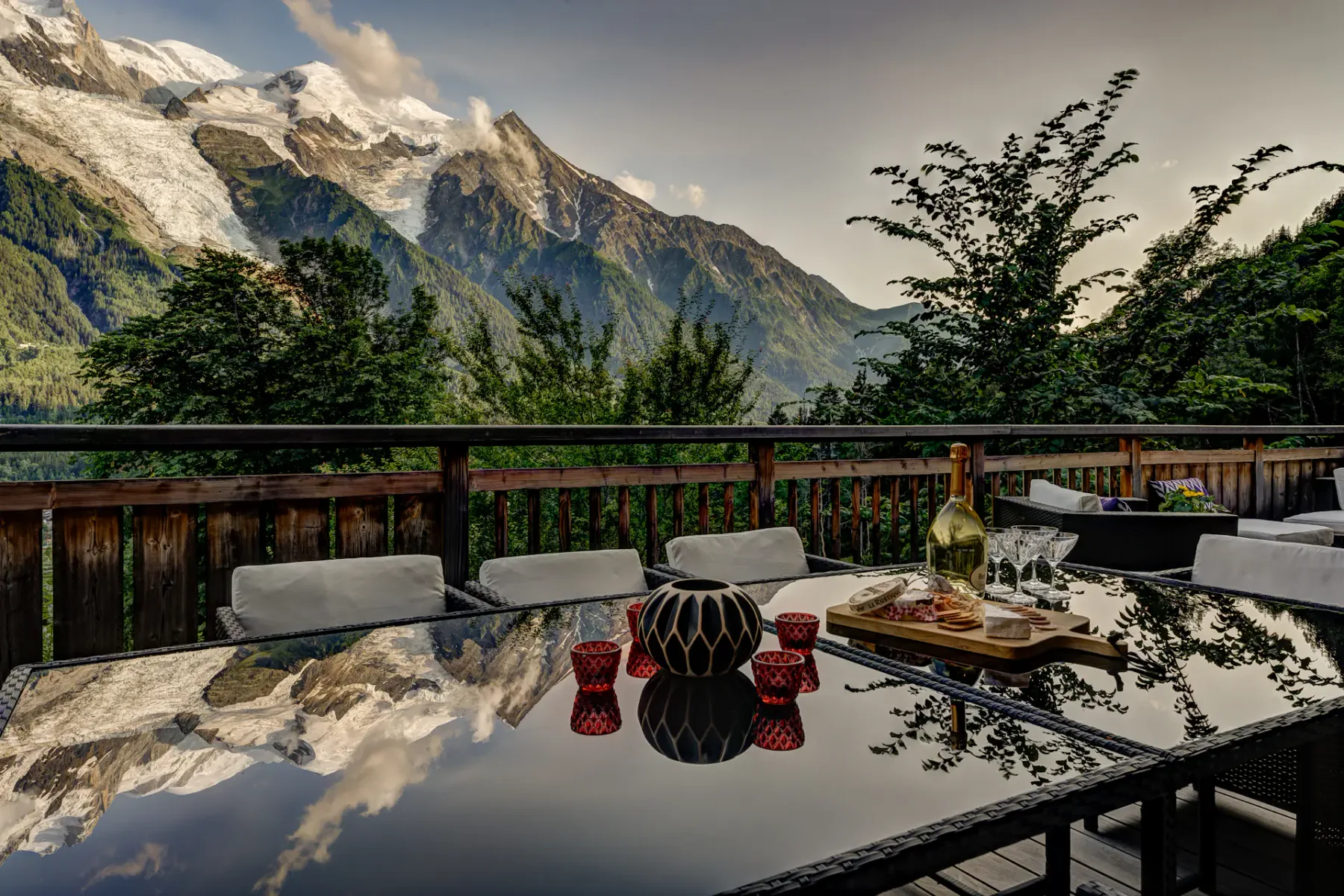 A reflective outdoor dining table overlooks a wooden terrace railing, green trees, and snow-capped mountains.