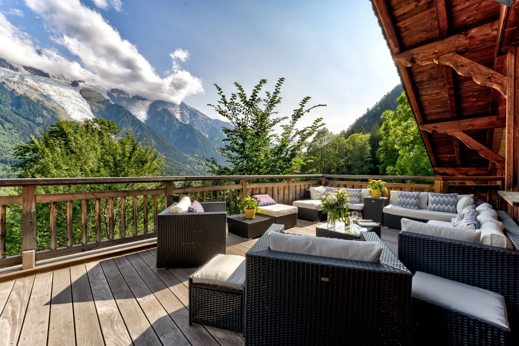 A wooden mountain-view terrace has wicker lounge seating, a glass-topped table, and a railing overlooking the peaks.