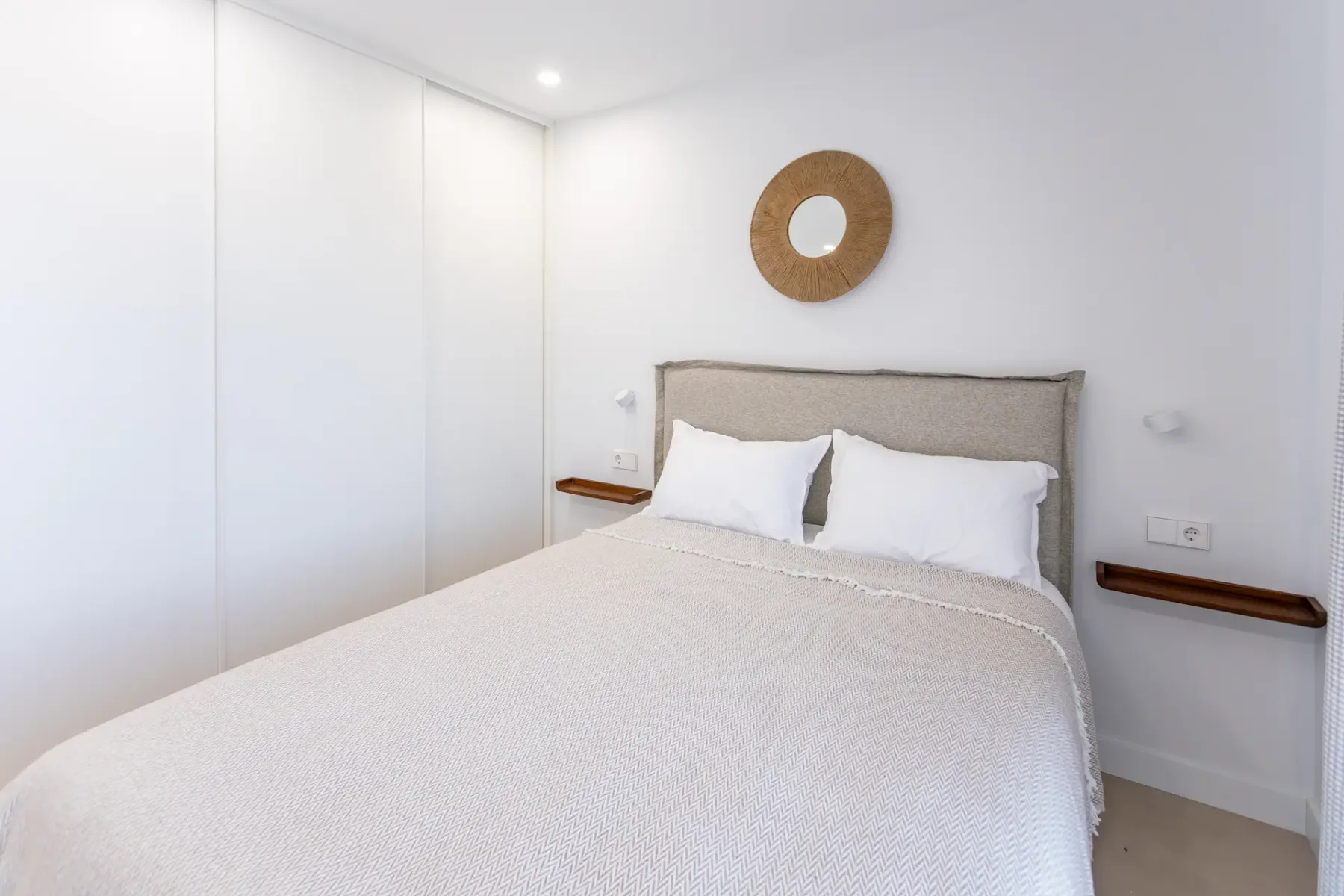 A bright bedroom with a neatly made bed and a round wall mirror.