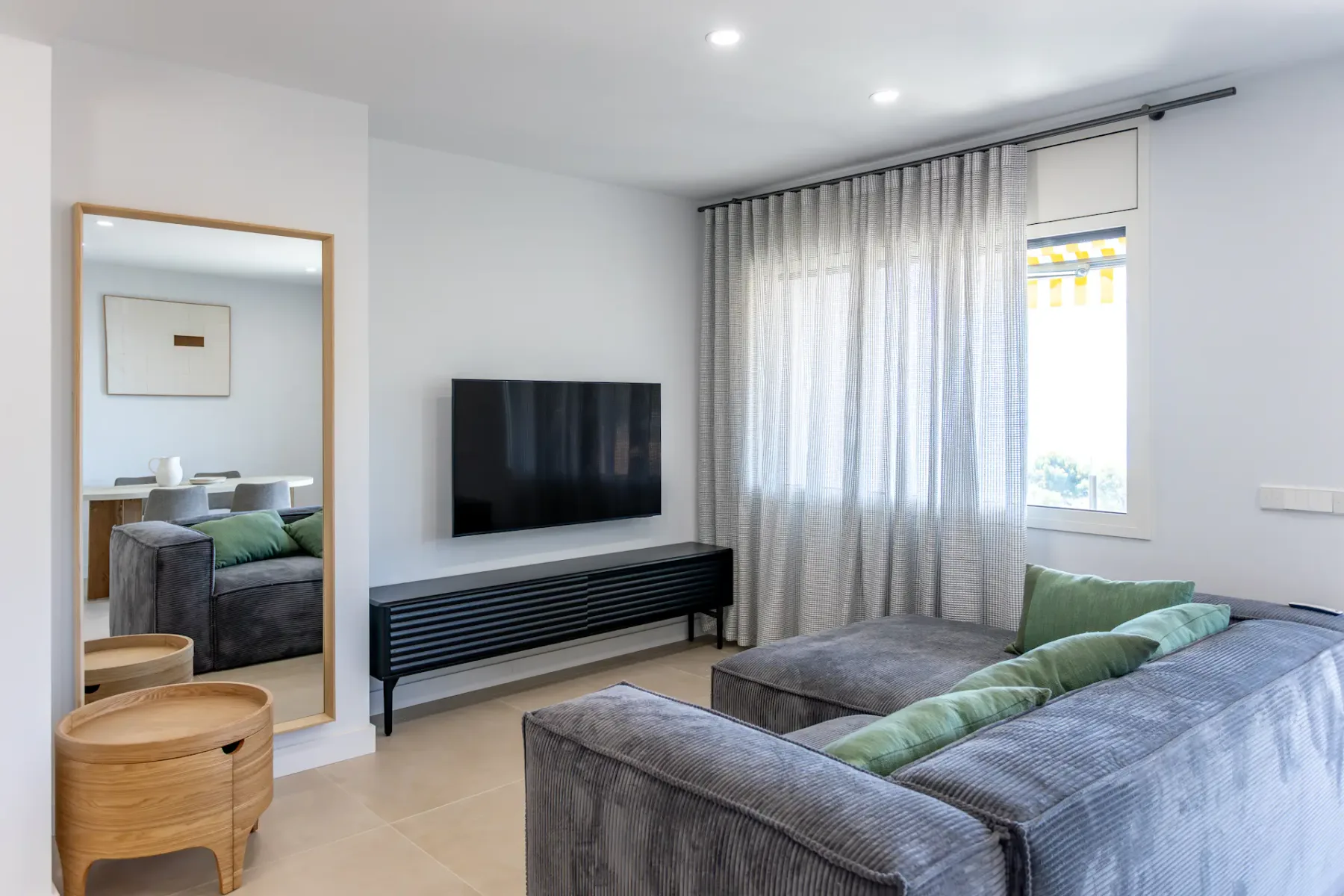 A living room with gray seating, a wall-mounted television, and curtains by the window.