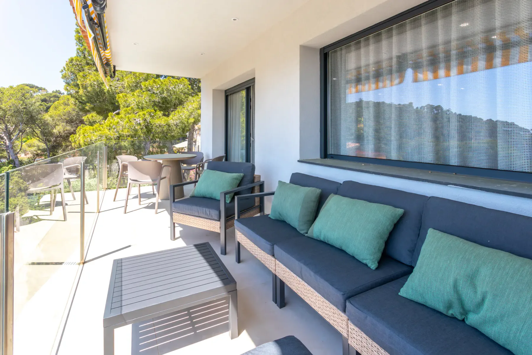 A covered patio has outdoor seating beside large windows and a glass railing.