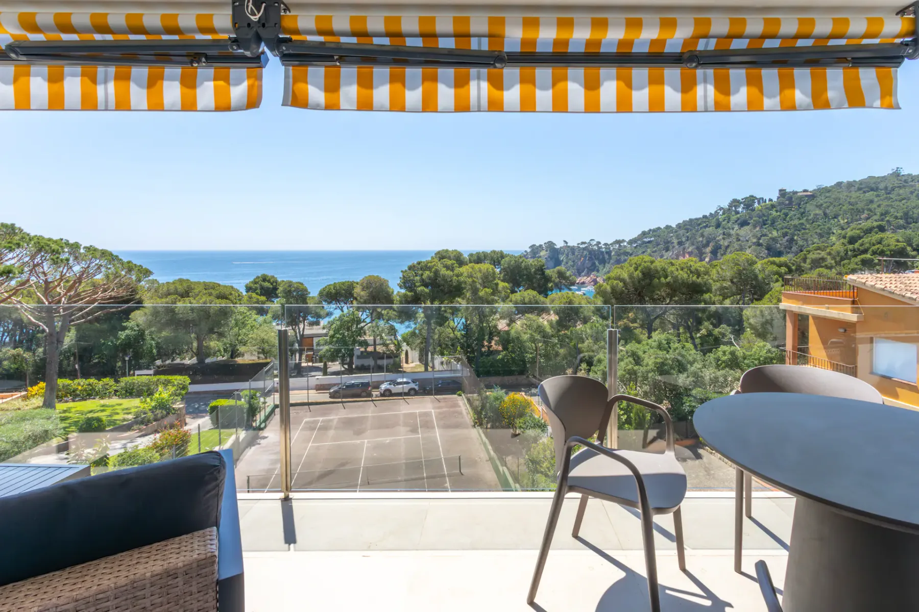 A balcony seating area overlooks a tennis court, green hills, and the sea.