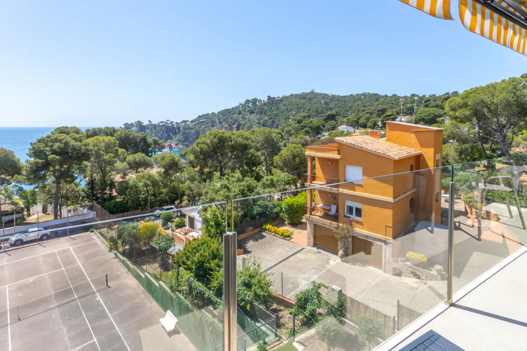 A sunny hillside view overlooks orange buildings, trees, and the sea.