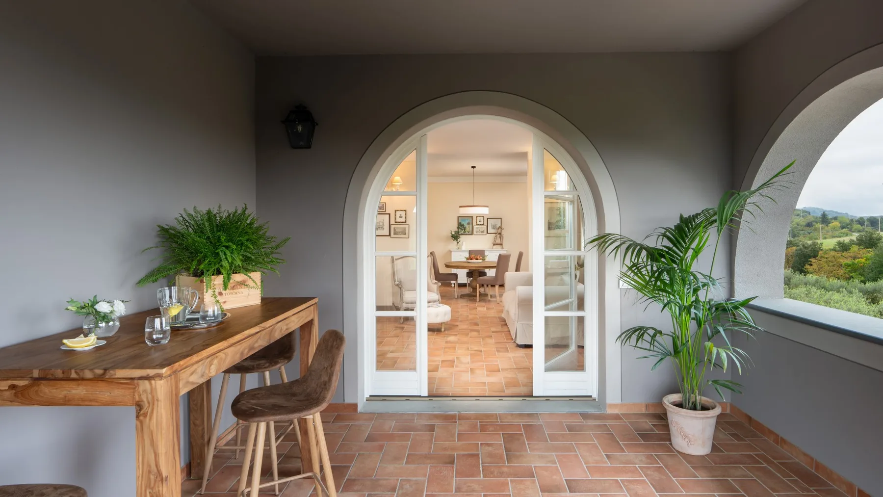 Un pasillo interior con suelo de baldosas da a un comedor por una puerta arqueada, con plantas y una mesa de madera.