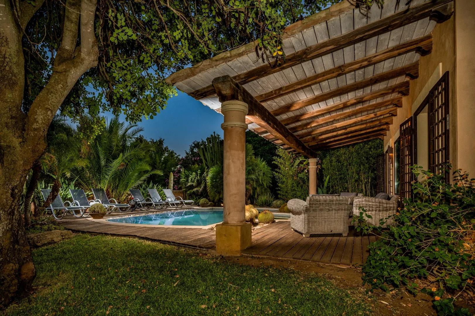 Un patio ajardinado junto a una piscina, con asientos bajo una terraza cubierta.