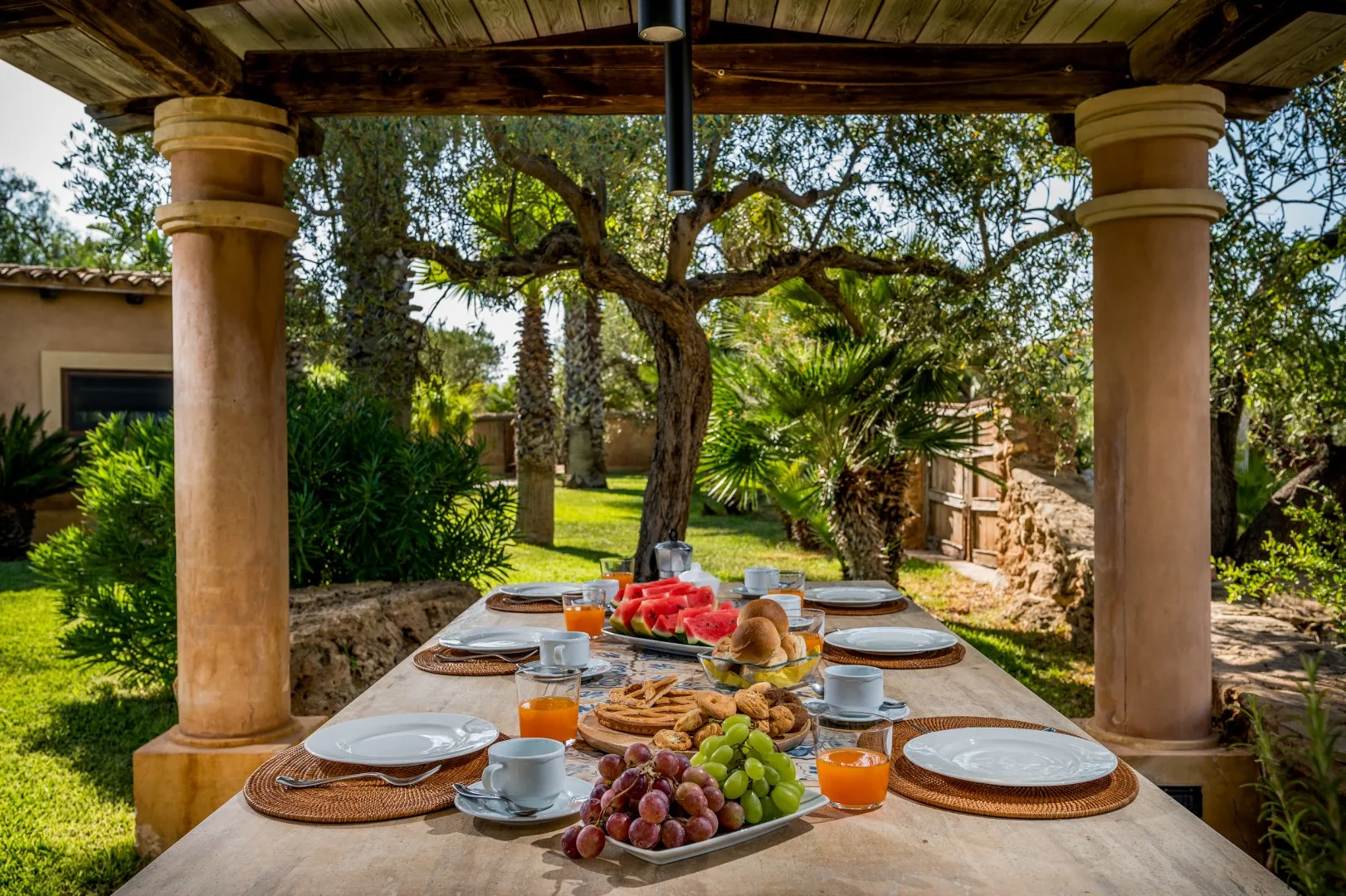Una mesa de comedor al aire libre está preparada con platos, fruta y desayuno bajo un patio cubierto.