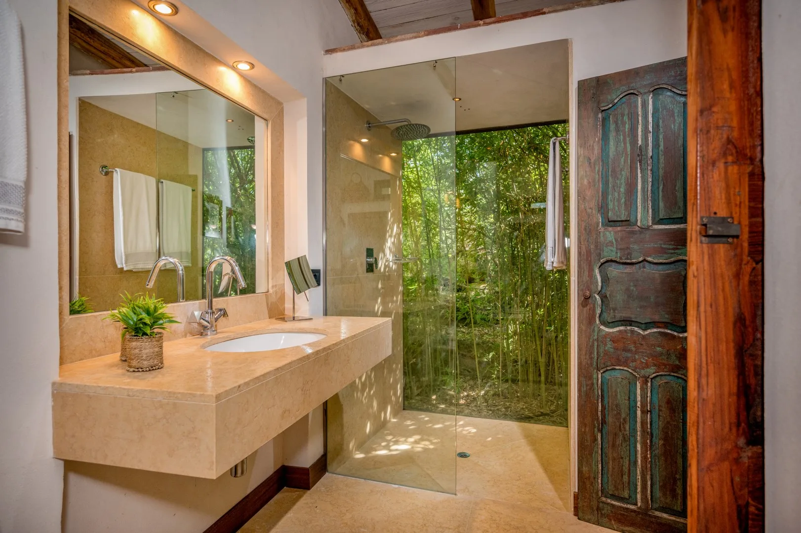 Un baño con lavabo de piedra, espejo y ducha de cristal frente a la vegetación.