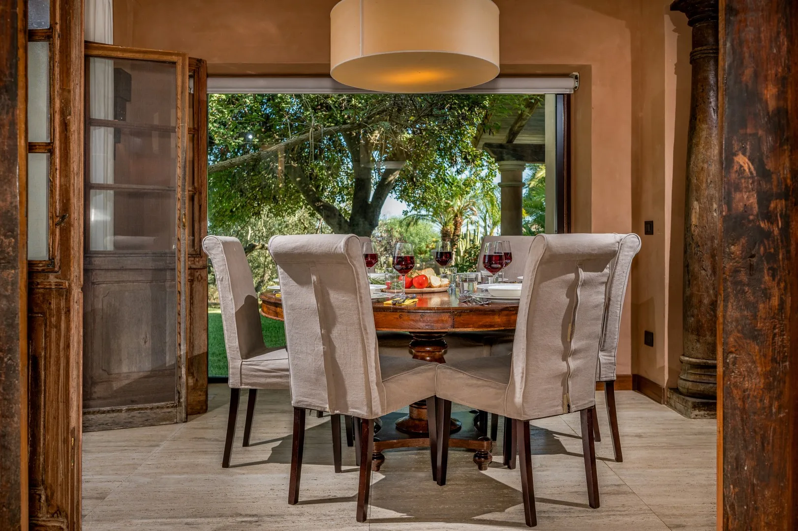 Un comedor con una mesa y asientos tapizados junto a puertas abiertas con vista a árboles.