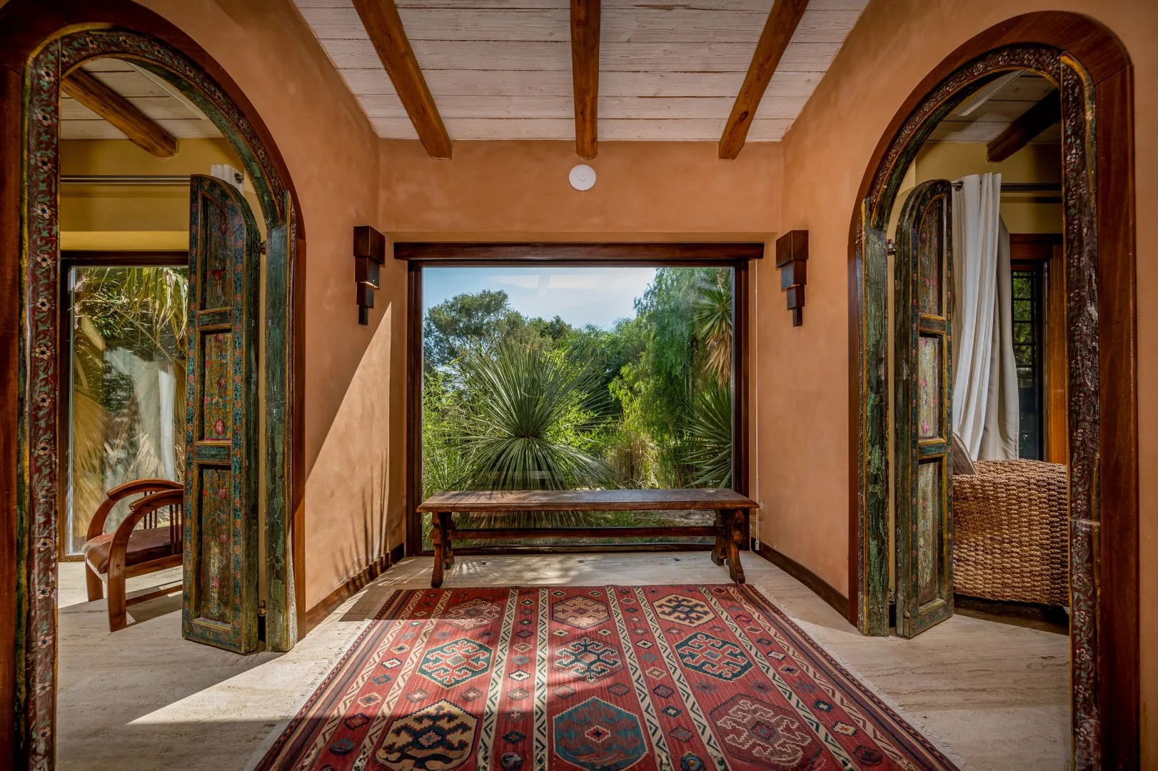 Una habitación de tonos cálidos se abre a un jardín frondoso, con una alfombra estampada en primer plano.