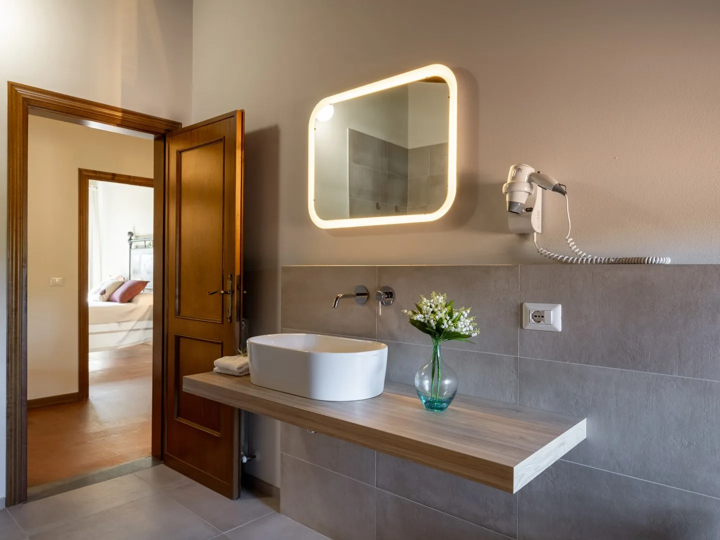 Mueble de baño con lavabo sobre encimera, espejo iluminado y una puerta de madera abierta.