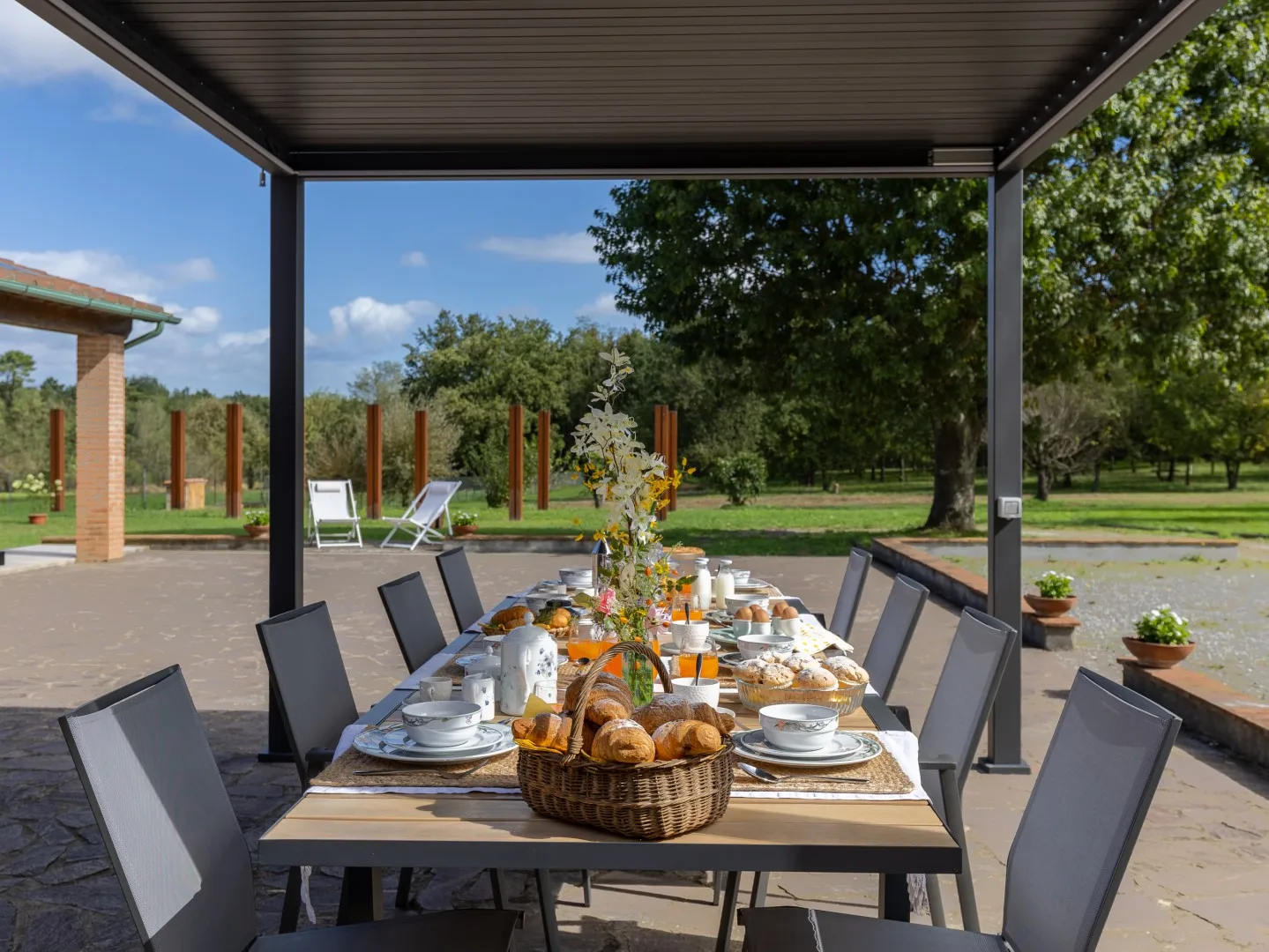 Una mesa está preparada bajo un patio cubierto al aire libre, con árboles y césped al fondo.