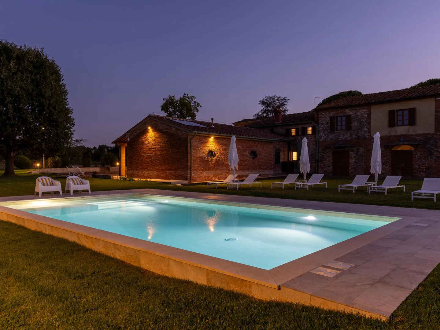 Piscina al aire libre junto a un edificio de ladrillo al anochecer, con asientos junto al borde.