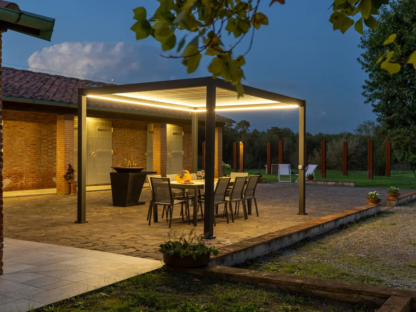 Un patio comedor al aire libre bajo una pérgola iluminada, con mesa y asientos.