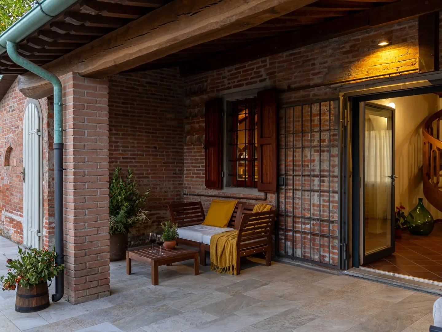Un patio cubierto de ladrillo tiene asientos de madera, una mesa pequeña y una puerta abierta.